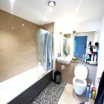 A beautiful modern London apartment available for sale by auction features a bathroom with tiled walls and floors, a bathtub on the left, and a toilet and sink on the right. Towels hang on a rail over the bathtub, while multiple toiletries are visible on the counter and adjacent shelves.