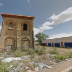 An old, abandoned brick building stands next to a modern, single-story structure with blue shutters. The surrounding area in Kent ME12 2PF is overgrown with weeds, and the sky above is partly cloudy. A brick wall with a metal gate marks the front of this Development Site Sheerness for sale by auction with World of Auctions.