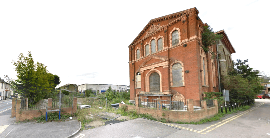 An abandoned, red-brick building with arched windows stands at a street corner in Sheerness, Kent ME12 2PF. Showing signs of decay with broken windows and overgrown vegetation, this development site features a tall, weathered tree on the left and is partially fenced off. For sale by auction with World of Auctions.