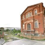 An abandoned, red-brick building with arched windows stands at a street corner in Sheerness, Kent ME12 2PF. Showing signs of decay with broken windows and overgrown vegetation, this development site features a tall, weathered tree on the left and is partially fenced off. For sale by auction with World of Auctions.