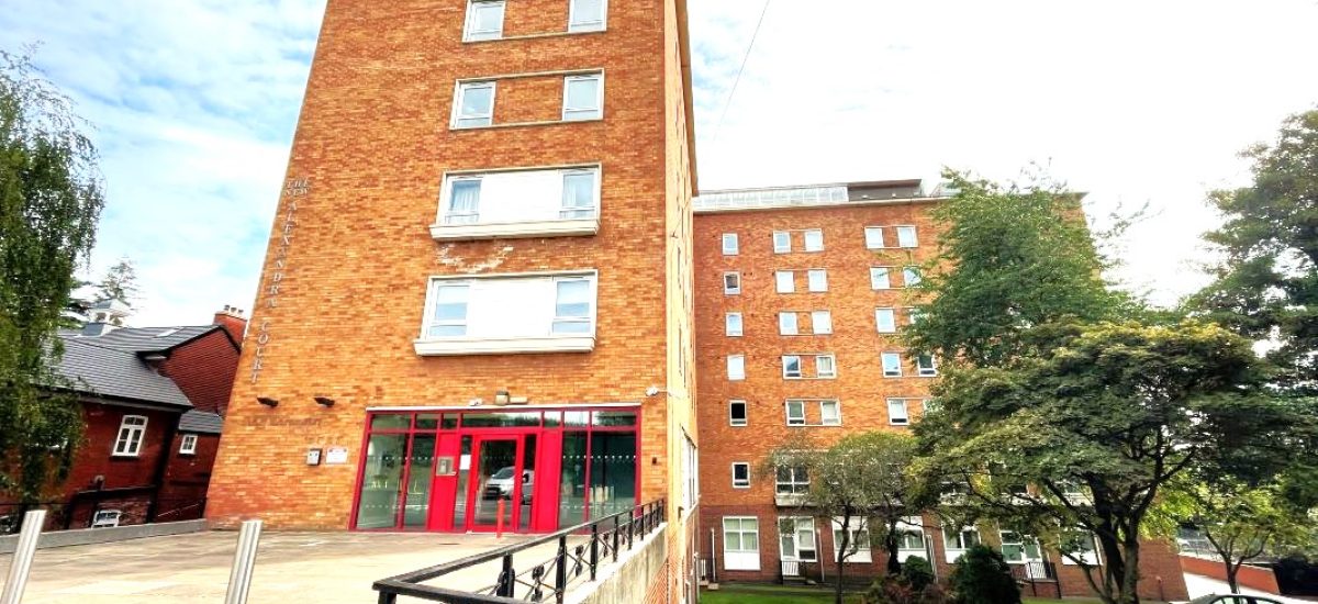A multi-story red-brick building with numerous windows and red-framed entrance doors stands proudly, surrounded by a neatly maintained lawn with trees and shrubs. Adjacent to the main building are smaller brick structures. The sky is partly cloudy—a picturesque view from New Alexander Court Nottingham, where World of Auctions has two investment apartments for sale by auction.