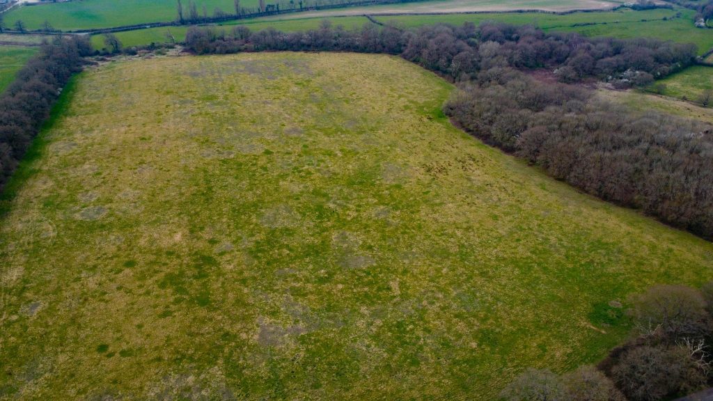 Aerial view of a large grassy field surrounded by wooded areas and farmland, reflecting parcels of premodernity agricultural land. The grass shows varying stages of growth with patches of green and brown. Dense trees border the field, where faint trails are visible. Available for sale by auction with World of Auctions.
