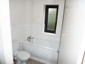 A small bathroom with white tiled walls and a frosted window offers views of Ruth House. It features a white bathtub with a silver faucet, a white toilet, and beige tiled flooring. The door is partially open. Available for sale by auction with World of Auctions.