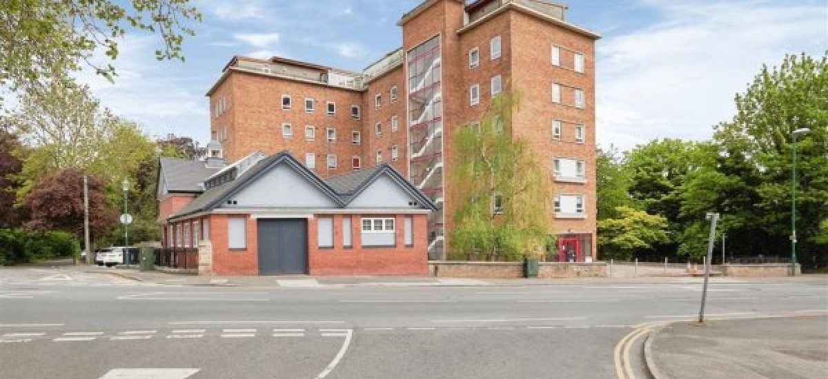 A large brick building with several stories on a corner lot, featuring a modern extension with a sloping roof. Surrounded by trees and greenery, New Alexandra Court Nottingham offers investment apartments for sale by auction through World of Auctions. The foreground shows road markings and a crosswalk on a clear, sunny day.