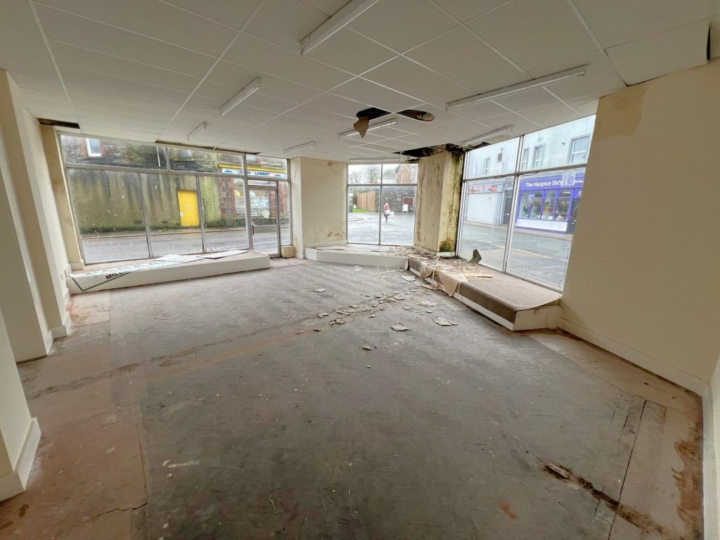 An empty, run-down storefront with large windows on 1-11 Wellington Street, Millom, Cumbria. The ceiling has a fan and fluorescent lights. The floor is covered with debris and the walls are peeling. This historic former co-op building offers a unique residential and commercial conversion opportunity for sale by auction with World of Auctions.