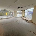 An empty, run-down storefront with large windows on 1-11 Wellington Street, Millom, Cumbria. The ceiling has a fan and fluorescent lights. The floor is covered with debris and the walls are peeling. This historic former co-op building offers a unique residential and commercial conversion opportunity for sale by auction with World of Auctions.