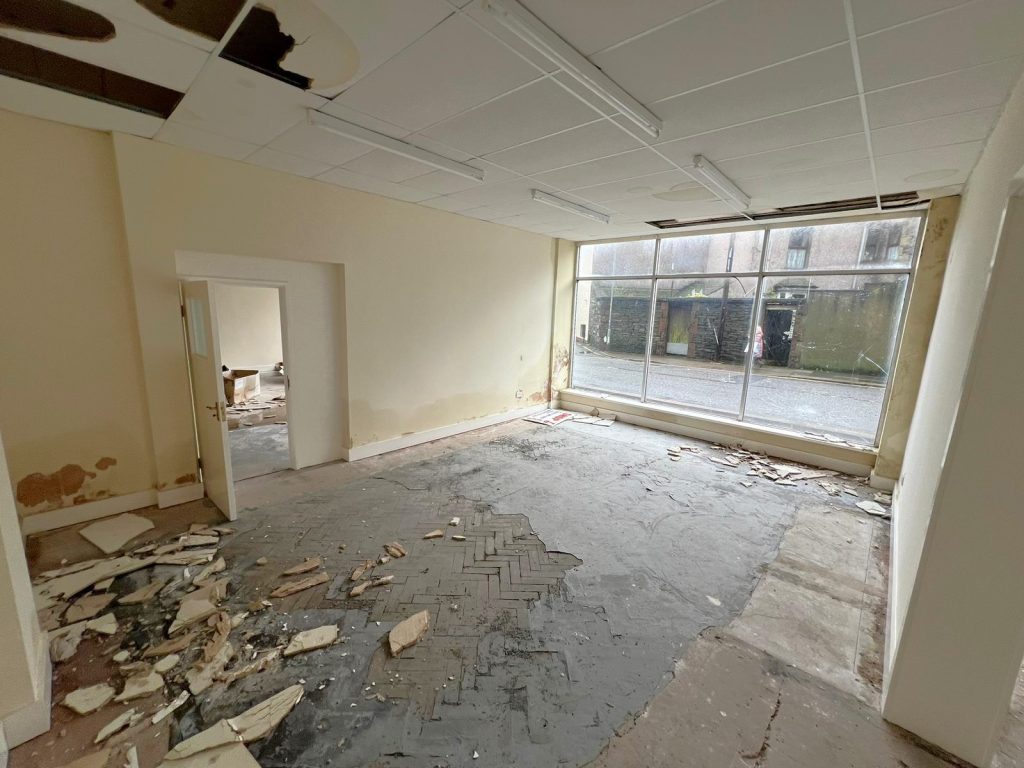 An abandoned, run-down room with broken ceiling tiles and debris scattered across the floor. The walls show signs of water damage and peeling paint. A large window at the far end reveals an empty, wet street outside. Located at 1-11 Wellington Street, Millom, Cumbria, this historic former co-op building offers a residential and commercial conversion development opportunity for sale by auction with World of Auctions