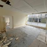 An abandoned, run-down room with broken ceiling tiles and debris scattered across the floor. The walls show signs of water damage and peeling paint. A large window at the far end reveals an empty, wet street outside. Located at 1-11 Wellington Street, Millom, Cumbria, this historic former co-op building offers a residential and commercial conversion development opportunity for sale by auction with World of Auctions