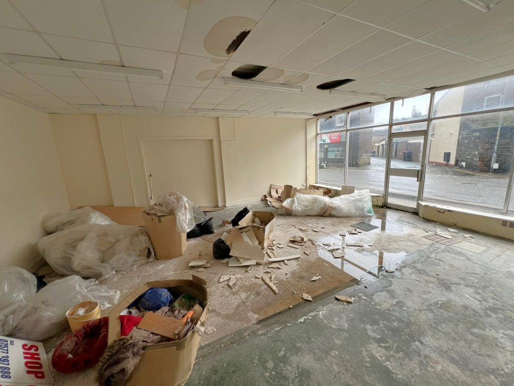 Empty room at 1-11 Wellington Street Millom, Cumbria, with a damaged ceiling, various debris, cardboard boxes, and large trash bags scattered on the floor. Large glass windows allow natural light in and offer a view of the street outside with neighboring buildings. Historic former co-op building - residential and commercial conversion development opportunity for sale by auction with World of Auctions.