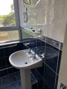 A bathroom sink with a blue and white tile backsplash. Above the sink, there is a small shelf with a mirror and a toothbrush holder mounted on the wall. A large window next to the sink offers a view of greenery outside. The floor is tiled in matching blue, complementing the quality in New Alexandra Court Nottingham's investment apartments for sale by auction.