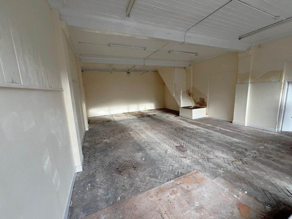 An empty room at 1-11 Wellington Street Millom, Cumbria, with cream-colored walls and a high white ceiling. The floor is made of dark, herringbone-patterned wooden planks. White ceiling lights illuminate areas where paint appears to be peeling. Natural light fills this historic former co-op building—an ideal residential and commercial conversion development opportunity for sale by auction with World of Auctions
