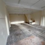An empty room at 1-11 Wellington Street Millom, Cumbria, with cream-colored walls and a high white ceiling. The floor is made of dark, herringbone-patterned wooden planks. White ceiling lights illuminate areas where paint appears to be peeling. Natural light fills this historic former co-op building—an ideal residential and commercial conversion development opportunity for sale by auction with World of Auctions