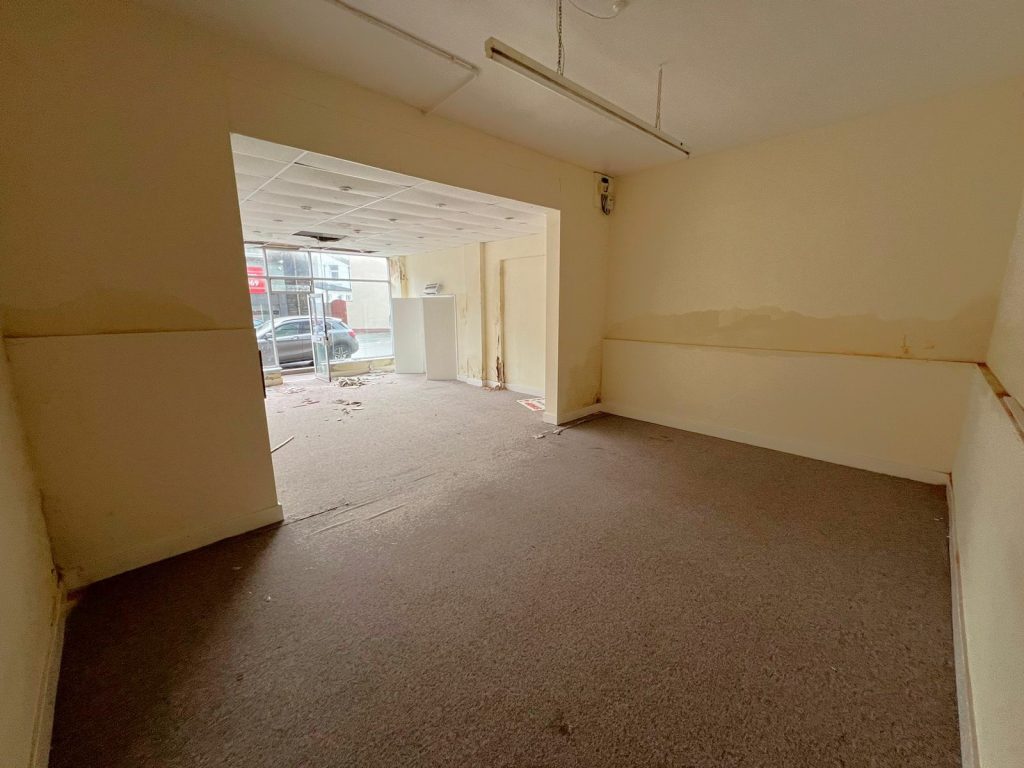 A vacant room at 1-11 Wellington Street, Millom, Cumbria, part of the historic former co-op building. This residential and commercial conversion development opportunity features beige walls and brown carpet flooring. Extending into another area via a doorway, large windows on the far side allow natural light to enter. Some cables and debris are visible on the floor. For sale by auction with World of Auctions