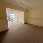 A vacant room at 1-11 Wellington Street, Millom, Cumbria, part of the historic former co-op building. This residential and commercial conversion development opportunity features beige walls and brown carpet flooring. Extending into another area via a doorway, large windows on the far side allow natural light to enter. Some cables and debris are visible on the floor. For sale by auction with World of Auctions