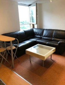 A cozy living room at New Alexandra Court Nottingham features a black leather L-shaped sofa positioned in the corner. In front of the sofa is a white square coffee table. An open window allows natural light to illuminate the space. Discover investment apartments for sale by auction with World of Auctions.