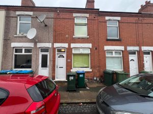 A row of terraced houses with brick facades, white doors, and windows. Two green trash bins are outside the house in the center. Red and grey cars are parked on a wet street. Satellite dishes adorn the houses. Ideal as CV2 4HQ, this high-yielding HMO investment property is up for grabs at World of Auctions, 59 Villiers Street, Coventry.