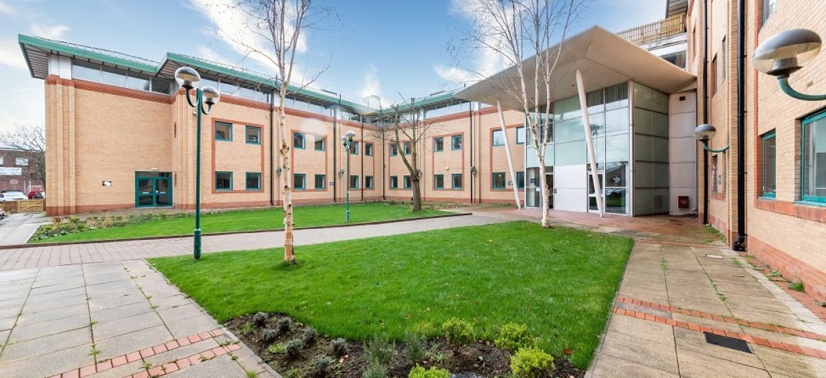 Humphry Davy House is a modern building with light brown brick walls and large windows, surrounding a courtyard with green grass, small trees, and a paved walkway. The sky is blue with wispy clouds. An entrance with a glass facade and angular roof is visible on the right. Investment flats are available for sale by auction below market value in this prime location from World of Auctions.