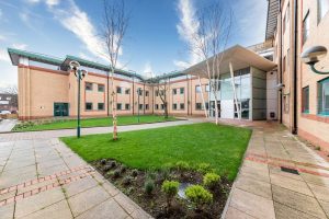 Humphry Davy House is a modern building with light brown brick walls and large windows, surrounding a courtyard with green grass, small trees, and a paved walkway. The sky is blue with wispy clouds. An entrance with a glass facade and angular roof is visible on the right. Investment flats are available for sale by auction below market value in this prime location from World of Auctions.