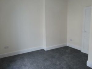 An empty room with light beige walls and dark gray carpet. There is a white door to the right, and a power outlet is visible on the left wall. The room has a simple, clean, and minimalistic appearance. Discover the potential of this space at 73 Blaen CF48 3RB, on the market with World of Auctions.