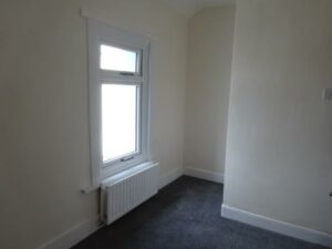 A small, empty room with white walls and a grey carpet. The room has a single window with a radiator below it. The window features white trim, and natural light is coming in, brightening the space. The room has a minimalist and clean appearance.