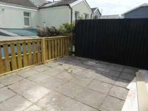 A small, paved patio area with a wooden fence on the left and a black, vertically slatted fence on the right. The patio is bordered by a house with light-colored walls. A few weeds are visible growing between the paving stones. Various materials lie in a corner at 73 Blaen CF48 3RB, on the market with World of Auctions.