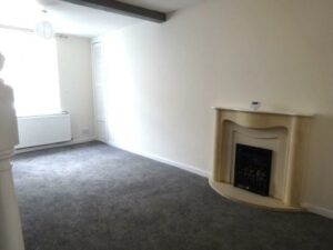 A minimalist living room at 73 Blaen CF48 3RB features a gray carpet and cream-colored walls. A white radiator sits beneath a window that lets in natural light. On the right wall, there's a beige fireplace with black accents. A small light fixture adorns the ceiling, adding subtle elegance.