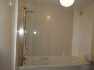 A modern bathroom with a white bathtub and a glass shower screen. There's a chrome shower head mounted on the wall above the tub. The walls are light-colored, and a round ceiling light is visible, illuminating the space. A partial view of a sink is on the right. Located at 73 Blaen cf48 3rb, now on the market with World of Auctions.