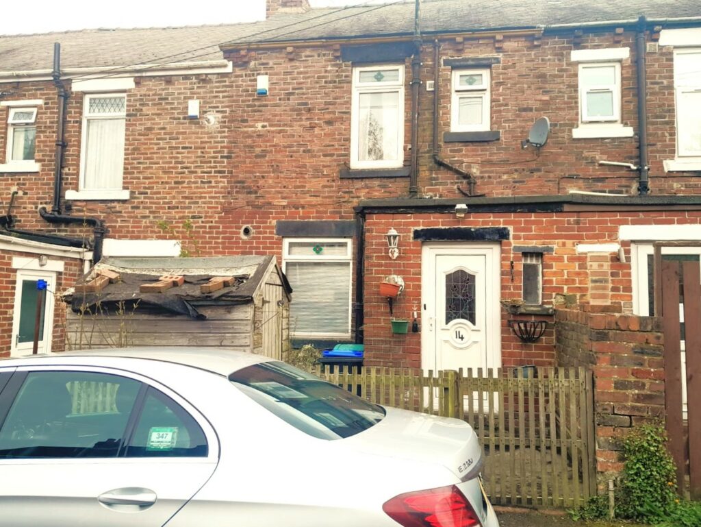 A brick townhouse at 14 Rose Avenue, Stanley, with a white door, small windows, and a picket fence in front. A silver car is parked outside on the street. The house has a small wooden shed and potted plants near the entrance. There is also a satellite dish on the wall. On the market with World of Auctions.