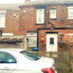 A brick townhouse at 14 Rose Avenue, Stanley, with a white door, small windows, and a picket fence in front. A silver car is parked outside on the street. The house has a small wooden shed and potted plants near the entrance. There is also a satellite dish on the wall. On the market with World of Auctions.