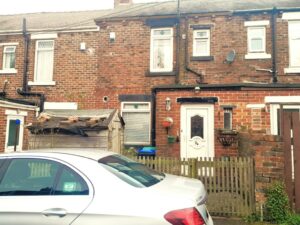 A brick townhouse at 14 Rose Avenue, Stanley, with a white door, small windows, and a picket fence in front. A silver car is parked outside on the street. The house has a small wooden shed and potted plants near the entrance. There is also a satellite dish on the wall. On the market with World of Auctions.