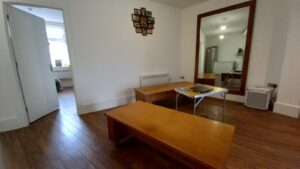 A minimalistic living room at 120 St Margaret's Road Twickenham features wooden flooring. It has two low wooden tables, one holding a laptop. The large wall mirror reflects a refrigerator and some kitchen space. There's a vent on the wall, a collage photo frame above it, and an open door to another room.