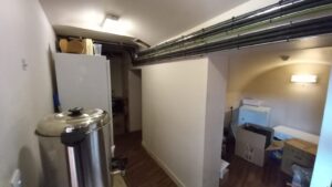 A narrow room with a slanted ceiling, featuring a large metal container on the left, possibly a water heater or boiler. The floor is wooden, and the room contains various boxes and items, including cabinets and shelves. The lighting is provided by wall-mounted fixtures.