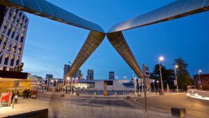 An urban scene at dusk features a modern sculpture of interlocking metal arches over a street. Surrounding the structure are tall buildings with illuminated windows, showcasing the lively atmosphere Coventry Property Investment Guide emphasizes. Pedestrians and cars are present, and streetlights illuminate the area.