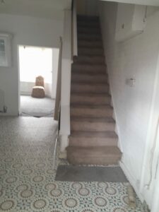 A narrow, carpeted staircase leads up from a small hallway with a patterned tile floor at 14 Rose Avenue. The hallway opens into a room with a large window, which illuminates a single armchair placed near it. The walls are painted white, and the space appears to be unfurnished. Now on the market with World of Auctions.
