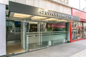 Property for sale by Auction A modern building entrance at Meridien House, 42 Upper Berkeley St., Marble Arch, London W1H 5QL features glass doors with silver accents, a covered walkway, and signage above the door. The adjacent red storefront, visible on the right, has floor-to-ceiling windows. A residential access sign is on the left side. Presented by World of Auctions