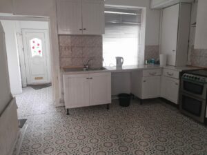 A kitchen with white cabinets, a sink, a kettle on the counter, and a black trash bin underneath. The floor has a patterned tile design, and a window with blinds is partially open. At 14 Rose Avenue Stanley, this home is newly on the market with World of Auctions.