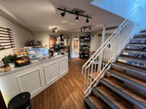 A cozy coffee shop at 120 St Margaret's Road, Twickenham, features an inviting interior with a counter displaying pastries, a coffee machine, and shelves with products. Wooden floors and white walls complement the staircase with white railings leading to an upper level. Track lighting creates a warm ambiance.