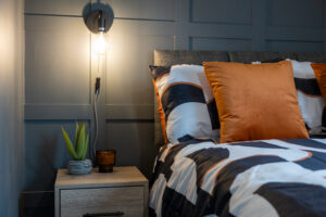 A cozy and modern bedroom features a bed with black, white, and orange patterned bedding, and two orange throw pillows. A small wooden nightstand next to the bed holds a potted plant and a candle. A wall-mounted lamp with a bare bulb illuminates the scene.