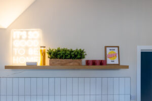 A wooden shelf mounted on a white tiled wall displays various decorations, including a neon sign reading "IT'S SO GOOD TO BE HOME," a plant in a wooden planter, three red mugs, and a framed print that says "THIS IS MY HAPPY PLACE.