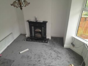 A room at 14 Rose Avenue, Stanley, with a grey carpet, white walls, and a black fireplace adorned with intricate designs. The room is lit by natural light streaming through a large window on the right. A chandelier hangs from the ceiling, and there are a few items and cables scattered on the floor. Now on the market with World of Auctions.