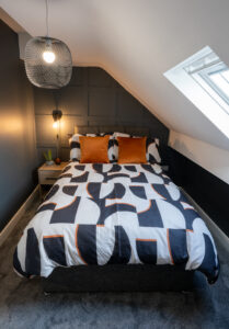 A cozy bedroom with a double bed covered in a modern black, white, and orange geometric patterned duvet. The room features an angled ceiling with a skylight, a bedside table with a lamp, a potted plant, and a gray and black color scheme.