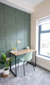 A minimalist home office features a light wooden desk with a black metal frame, a light green chair, a white desk lamp, and a pot with pencils. A plant in a white pot sits on the floor to the left. The wall behind is green with a grid pattern, and a large window is on the right.