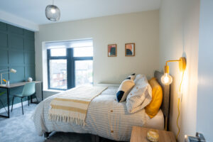 A cozy bedroom with a neatly made bed featuring striped bedding, yellow and black accent pillows, and a yellow throw. There is a small bedside table with a jar and a hanging exposed bulb lamp. A window offers natural light, and a desk with a chair is by the opposite wall.