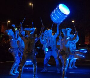 A group of performers dressed in reflective, futuristic costumes illuminated by blue LED lights playing drums at night. One performer is lifting a large illuminated drum above their head while others play smaller drums. In the backdrop, an ad for Leeds Property investment advice subtly contrasts the bright attire.