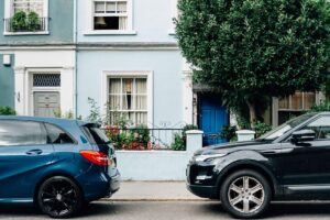 Two cars are parked on a street in front of a light blue townhouse with a blue front door. One is a blue hatchback, the other a black SUV. There is a large tree and some greenery in front of the house, and a window with an open shutter on the first floor. Why sell through World of Auctions? That’s easy! It's quick!