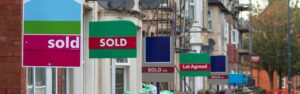 A row of houses with different colored real estate signs displaying "SOLD" and "Let Agreed" posted in front. Joint agents and professional service Providers manage these homes, lined with satellite dishes in a bustling urban neighborhood. Trees and a street are visible in the background.