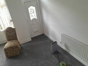 A simple room at 14 Rose Avenue, Stanley, with a white door featuring a floral design on its window. A patterned chair sits in the left corner. A radiator lines the right wall, and a portable heater with a green cord rests on the grey carpeted floor. On the market with World of Auctions.