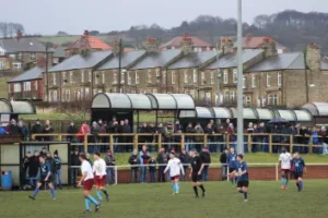 Property for sale by Auction A local soccer match is taking place on a grassy field with players in various colored uniforms. Spectators, some under covered stands, are watching the game. In the background, there are rows of houses with chimneys, and leafless trees can be seen. Presented by World of Auctions