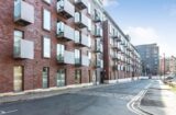 Property for sale by Auction A modern, multi-story brick apartment building with numerous balconies and large windows. The building lines a clean, empty street that curves slightly. Another building is visible in the background under a clear blue sky. Presented by World of Auctions