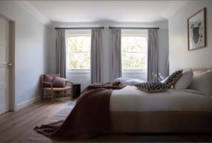 A neatly arranged bedroom features a large bed with white linens and a brown throw. Two windows with grey curtains allow natural light to stream in. A wooden chair with cushions is in the corner, and a piece of artwork hangs on the wall. The room has a serene ambiance.